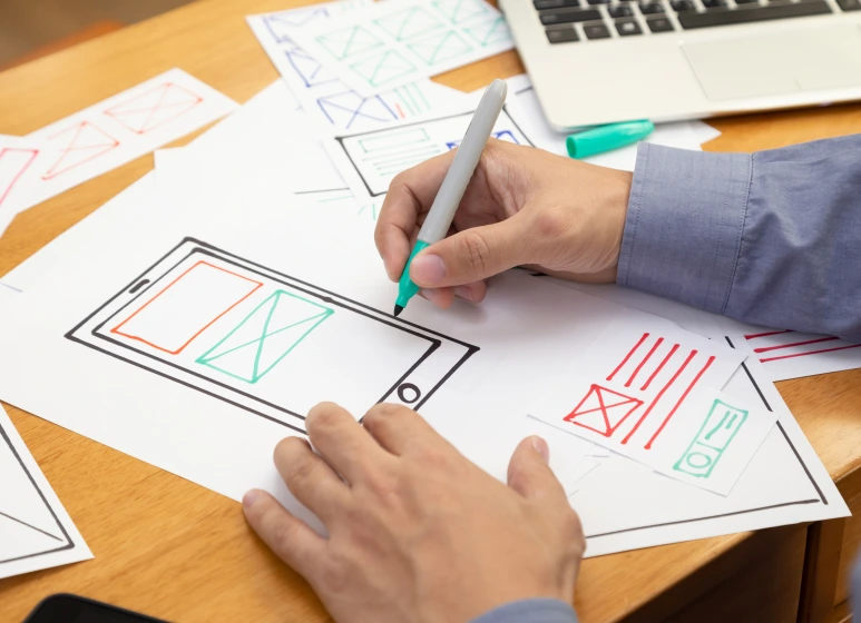 Person Sketching Mobile Interface Wireframes: A close-up of hands drawing mobile app interface wireframes on paper, symbolizing the design process for user-centric and visually appealing digital interfaces.
